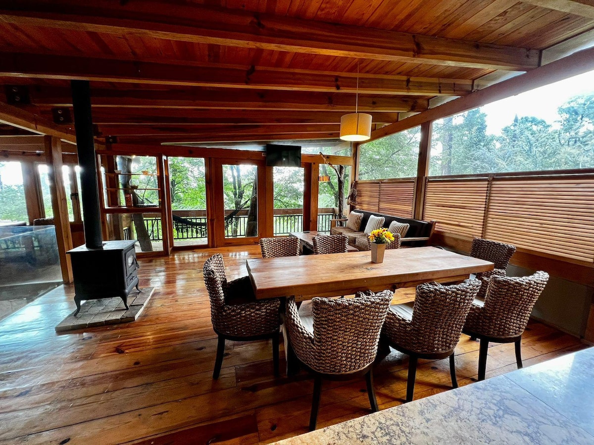 An inviting dining area features a long wooden table surrounded by woven chairs. Large windows provide a view of the trees outside, and a small wood stove is positioned nearby. Natural wood beams and floors create a warm environment.