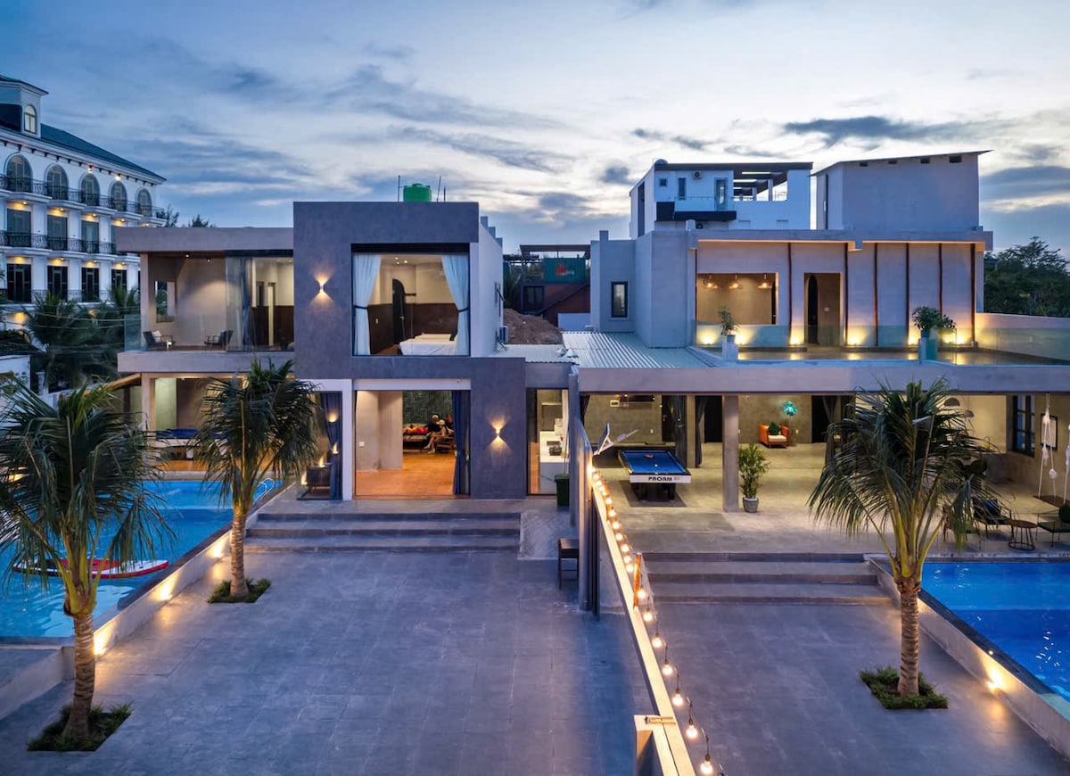 An elegant villa complex is displayed at dusk, featuring modern architecture with expansive outdoor areas. Two swimming pools are visible, bordered by palm trees and well-lit pathways. The open design allows for views of the interior spaces, showcasing an inviting ambiance.