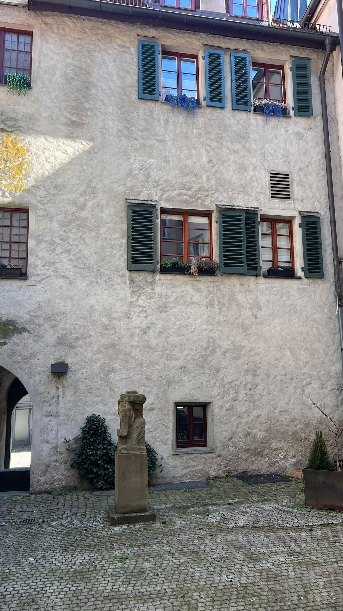 A textured exterior wall of a historic building is displayed, featuring dark green shutters on the windows. Decorative flower pots can be seen along the windowsills. A stone statue is positioned in the courtyard, surrounded by cobblestone paving and greenery.