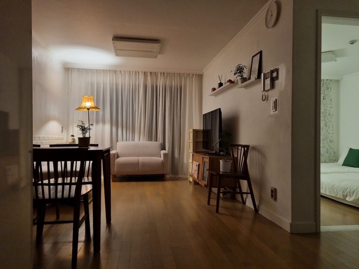 A well-lit living area features a simple dining table with four chairs beside a cozy sofa. Light-colored curtains soften the room while providing privacy. A television is positioned on a shelf against the wall, enhancing the home's functional design.