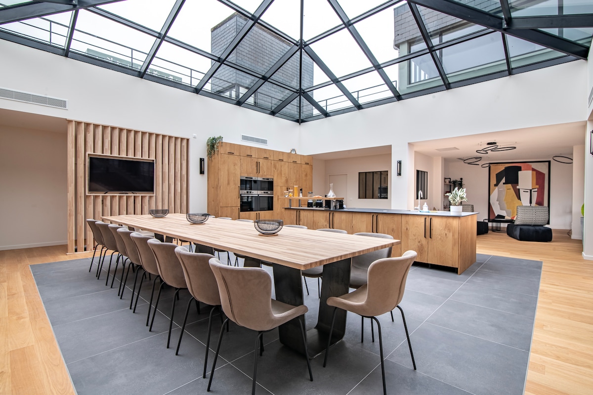 A spacious dining area features a long wooden table surrounded by several upholstered chairs. A sleek kitchen is visible in the background, complemented by wooden cabinetry and modern appliances. Large glass panels allow natural light to flood the room, enhancing the open and airy feeling.