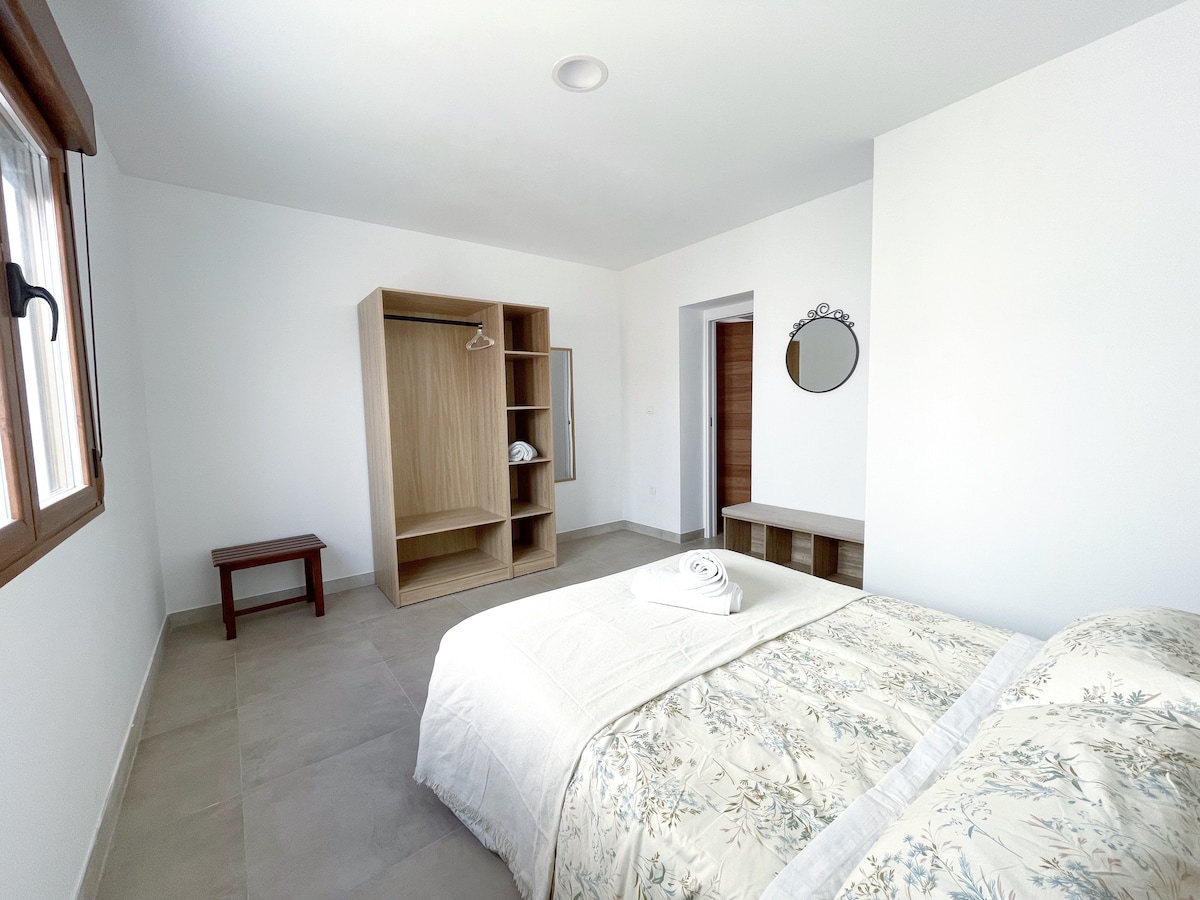 A spacious bedroom features a double bed covered with a light floral bedspread. A wooden bench and a small table are positioned nearby. A wall-mounted mirror reflects light, while a wardrobe with shelves stands adjacent. Large windows allow natural light into the room.