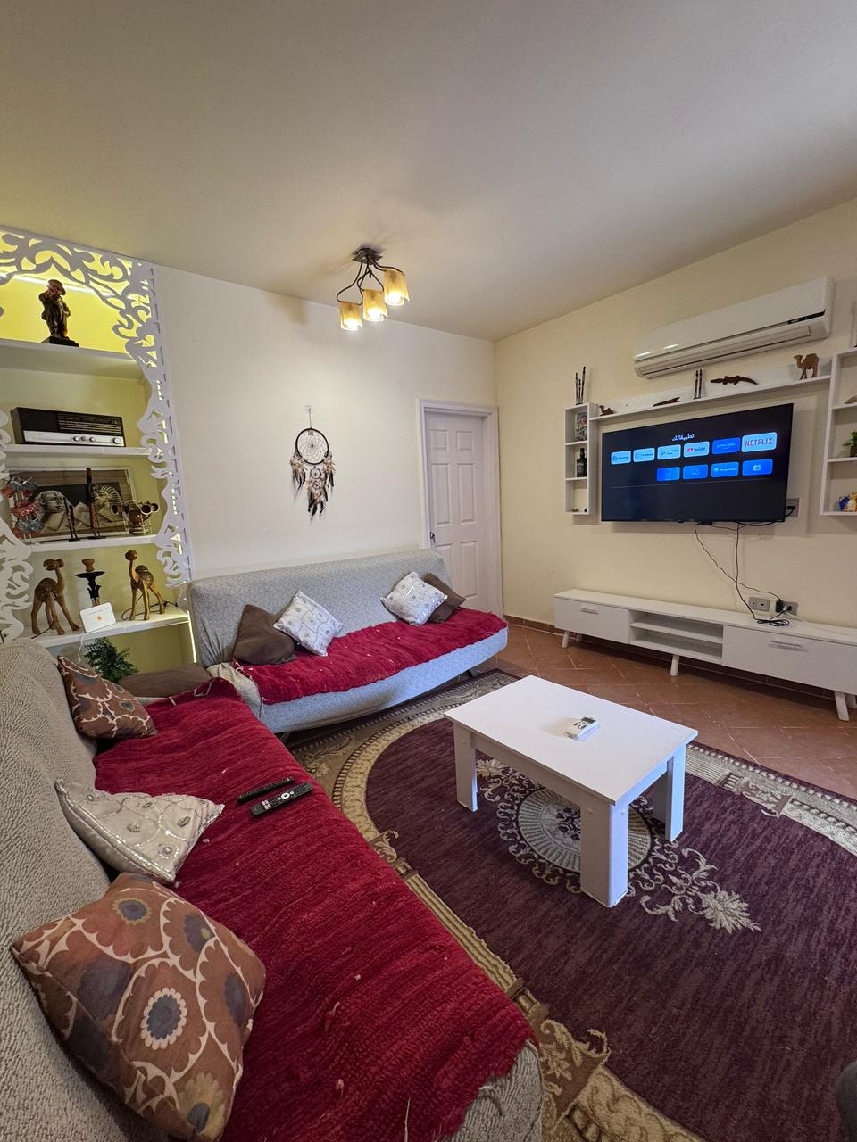 A welcoming living area is showcased, featuring two gray sofas adorned with red and white cushions. A white coffee table is centered on a patterned rug. A wall-mounted television is visible, accompanied by shelves displaying decorative items. Soft lighting adds to the relaxed ambiance.
