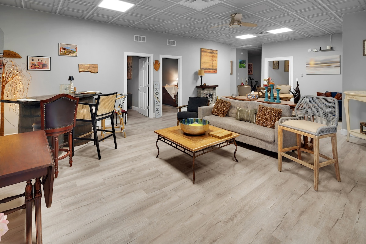 The living area features an open layout with stylish furniture, including a cozy sofa and assorted seating options. Decorative elements and artwork are placed throughout, enhancing the room's character. Natural light filters in, highlighting the wooden flooring and spacious design.