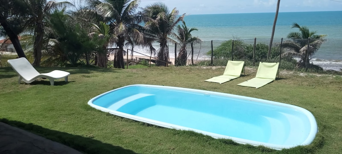 A serene pool is set within a grassy area, surrounded by palm trees and overlooking the calm ocean. Two lounge chairs are positioned near the water's edge, inviting relaxation. The beach is visible beyond a simple fence, with gentle waves lapping at the shore.