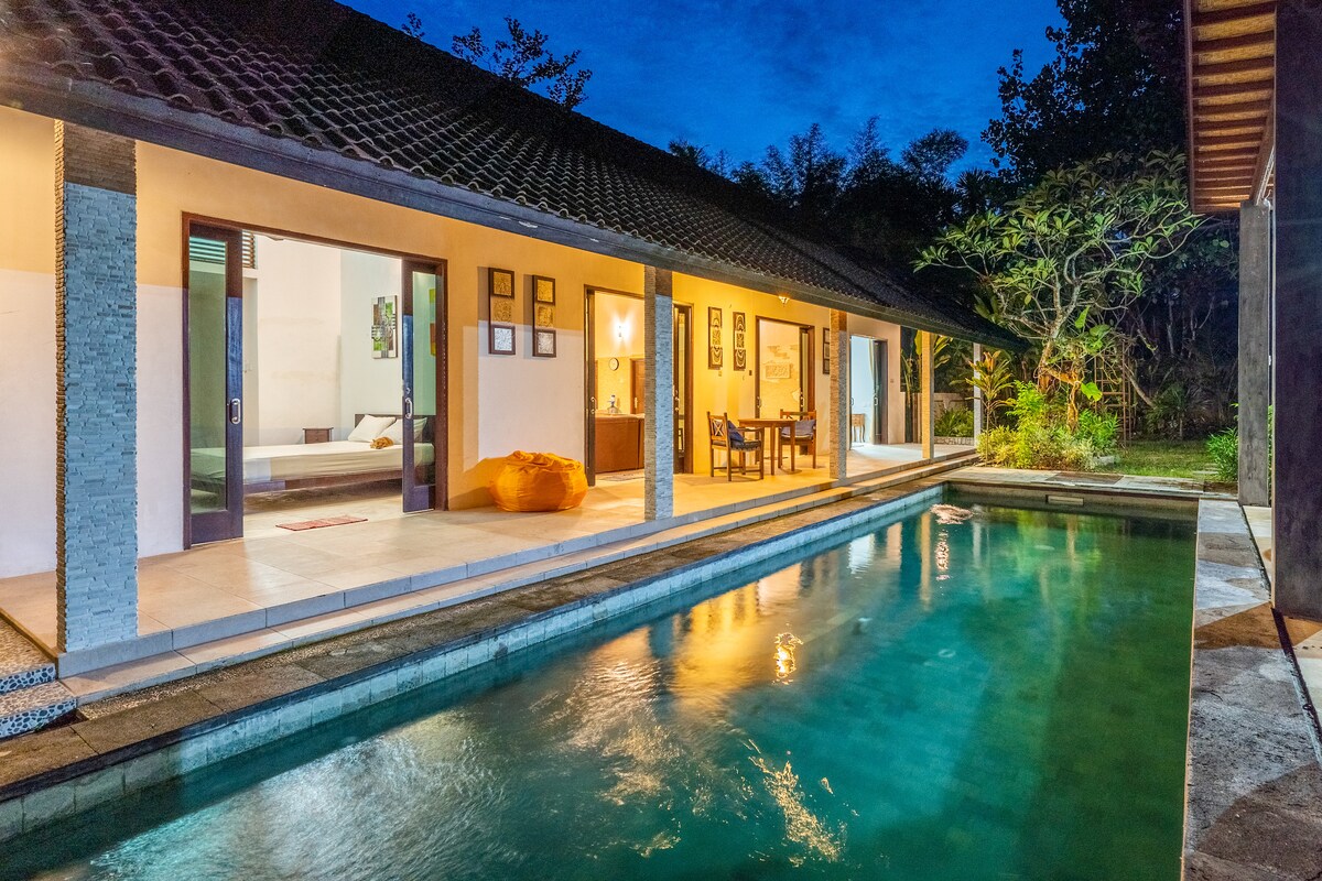 The image shows a serene villa with a private pool, reflecting ambient light from the evening sky. A spacious outdoor area is framed by large glass doors leading into the living space. Lush greenery surrounds the property, enhancing the tranquil environment.