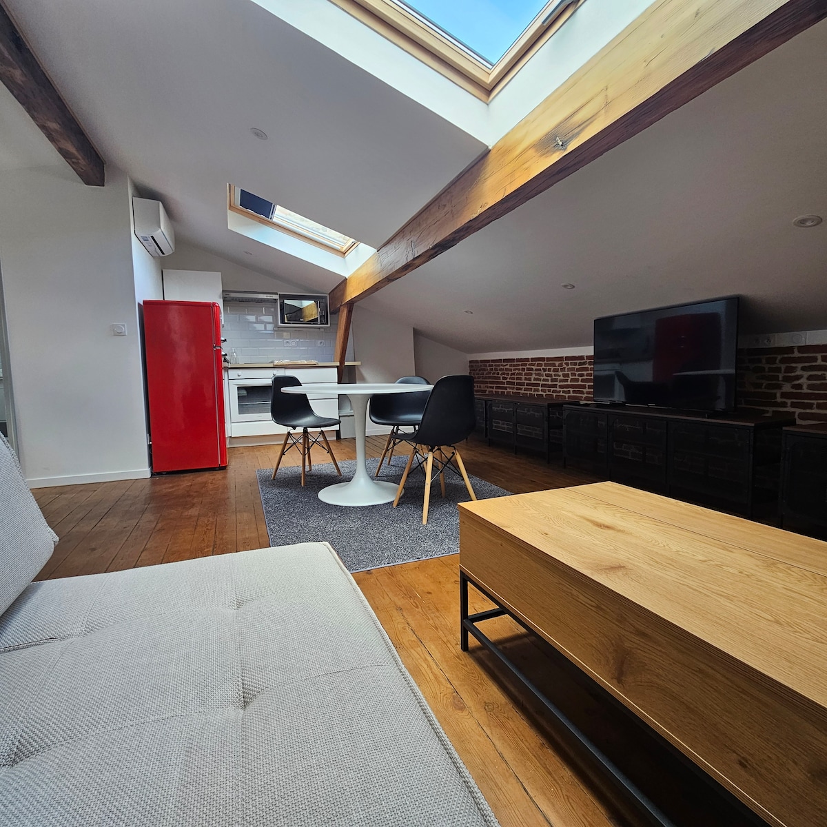 An open-concept living space is highlighted with a red refrigerator and a sleek white table surrounded by black chairs. Natural light floods in through a skylight, illuminating the wooden beams and warm-toned flooring. A large TV is positioned across from a gray sofa and wooden coffee table.
