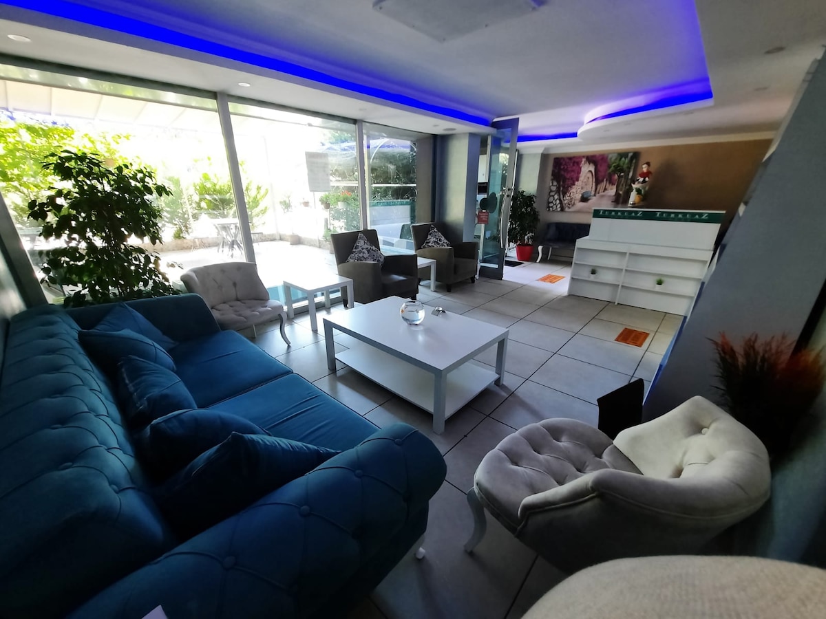 A modern lounge area features a plush blue sofa accompanied by light-colored armchairs. A white coffee table sits in the center, surrounded by lush greenery visible through expansive glass windows. Soft lighting creates a relaxed environment.