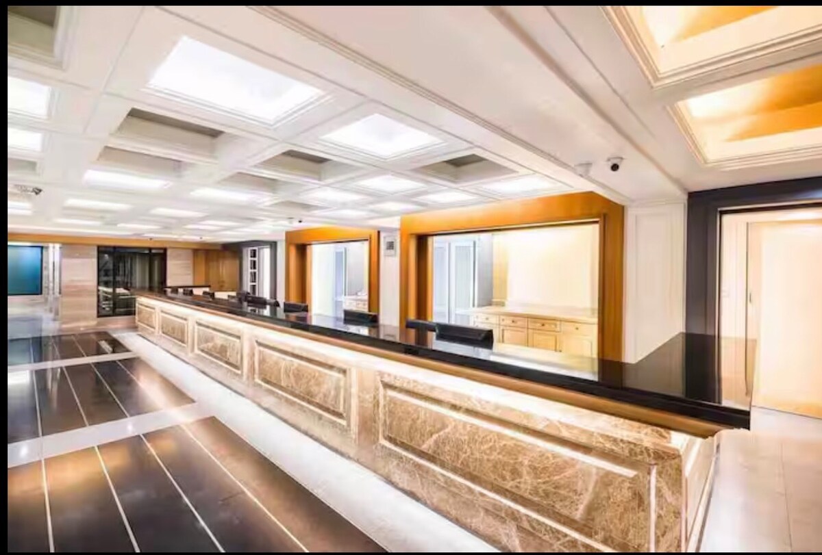 A spacious reception area is highlighted by a long, polished counter topped with marble. Brightly lit ceilings enhance the modern design, featuring a mix of recessed lights and decorative panels. Large windows provide a view into the adjoining spaces, creating an open and inviting atmosphere.