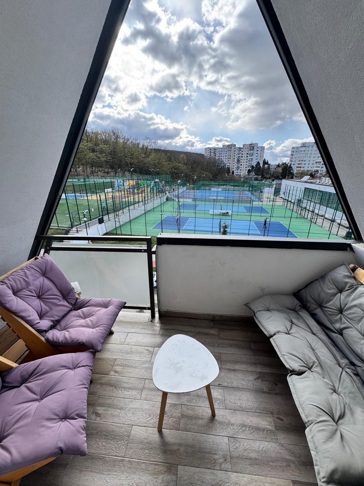A balcony area is visible, featuring cozy seating arrangements with two cushioned chairs and a small side table. The view reveals multiple tennis courts below, set against a backdrop of trees and nearby buildings, under a partly cloudy sky.