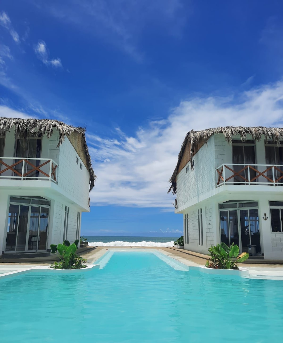 Two charming structures with thatched roofs are situated on either side of a tranquil swimming pool. Lush plants flank the pool area, while an expansive ocean view is visible beyond the pool. Bright blue skies and soft clouds frame the scene.