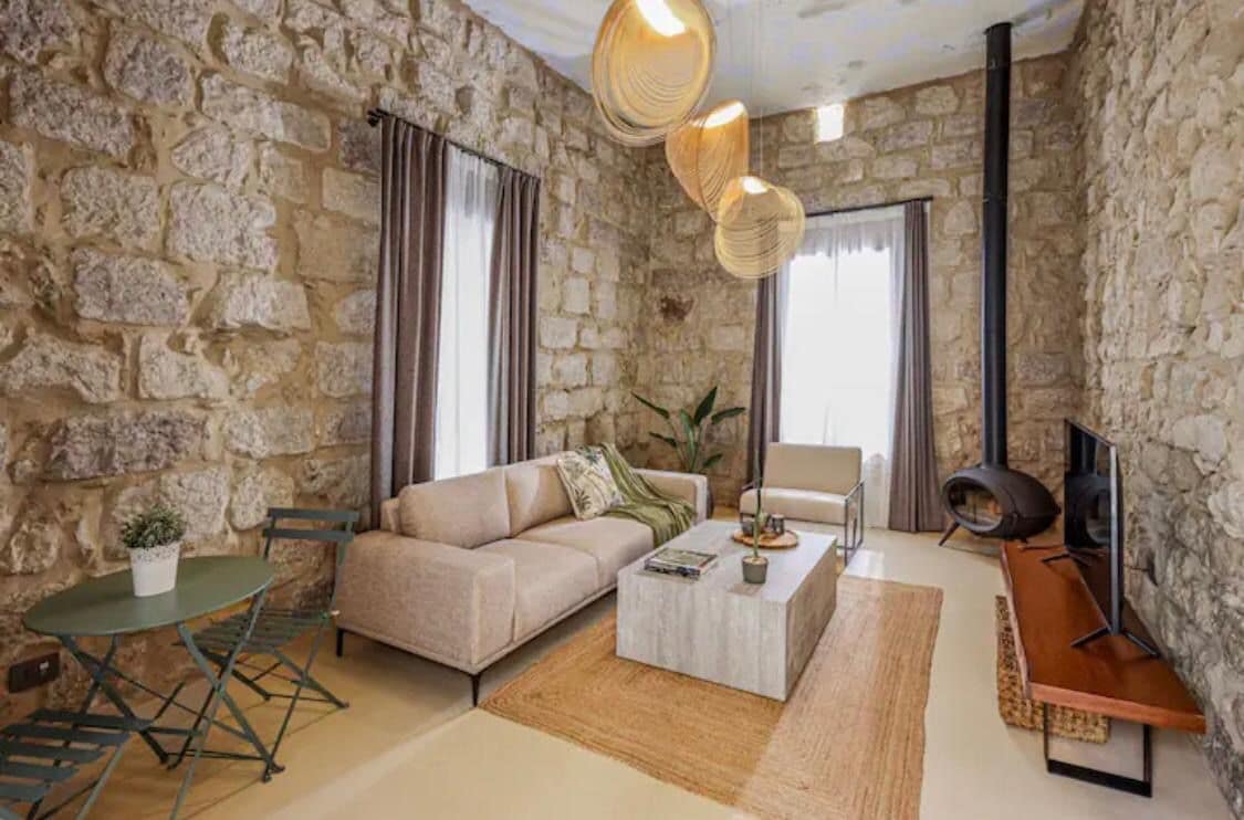 A living room features textured stone walls and soft lighting from pendant fixtures. A light-colored sofa and an armchair are arranged around a rectangular coffee table. A small green table with folding chairs is present, and a sleek television is positioned on a wooden console.