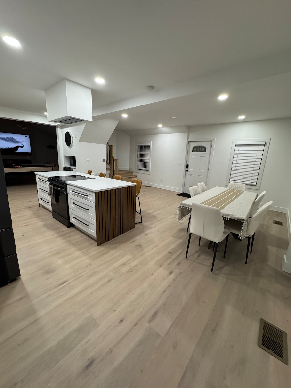 A modern kitchen and dining area is featured, showcasing an island with a stovetop and ample cabinetry. A spacious dining table is surrounded by light-colored chairs. Bright lighting enhances the clean aesthetics, with a doorway leading to an exterior patio visible in the background.