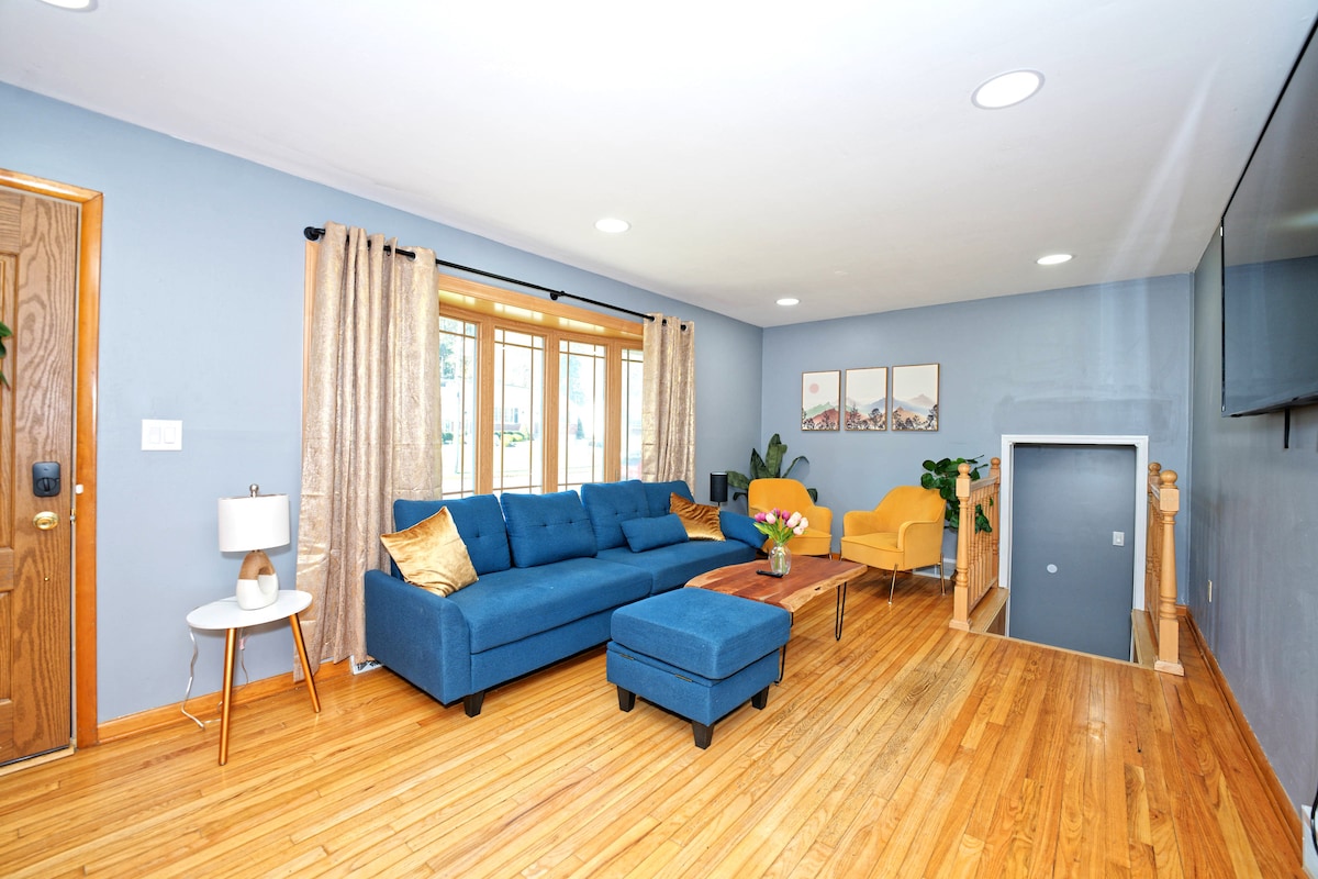 A spacious living area features a deep blue sofa paired with a yellow chair. A wooden coffee table sits in the center, adorned with a vase of flowers. Large windows provide natural light, and soft curtains frame the view. Warm wood flooring complements the overall design.