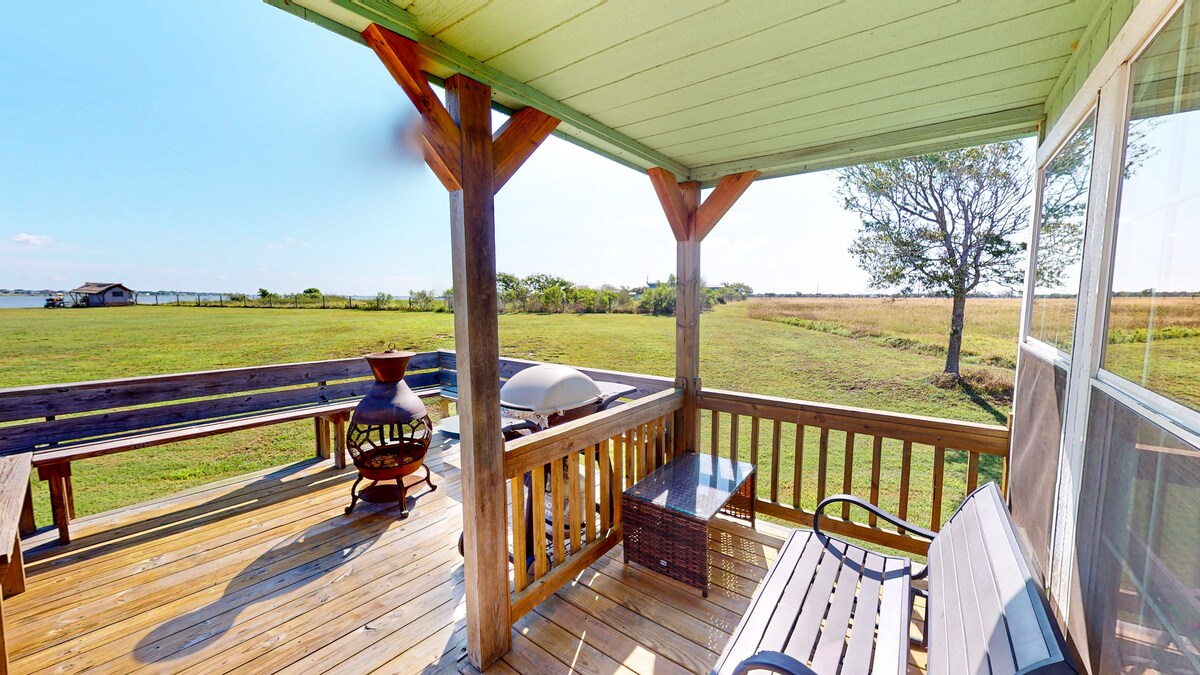 A spacious porch is shown with a wooden railing and seating areas. The floor is made of light-colored wood. A table and a fire pit are positioned for relaxation. Open greenery and a water view extend into the distance, creating a serene outdoor setting.