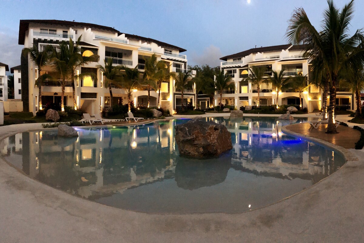 A serene pool area is surrounded by palm trees and stylish apartment buildings. The water reflects soft lighting, with a prominent rock feature in the center. Lounge chairs are positioned along the pool's edge, creating a welcoming outdoor space for relaxation.