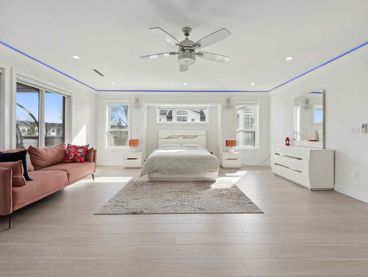 A spacious bedroom features a large bed flanked by modern bedside tables with lamps. Natural light filters through multiple large windows, illuminating a stylish seating area with a faded area rug. A ceiling fan adds a touch of modernity to the airy space.