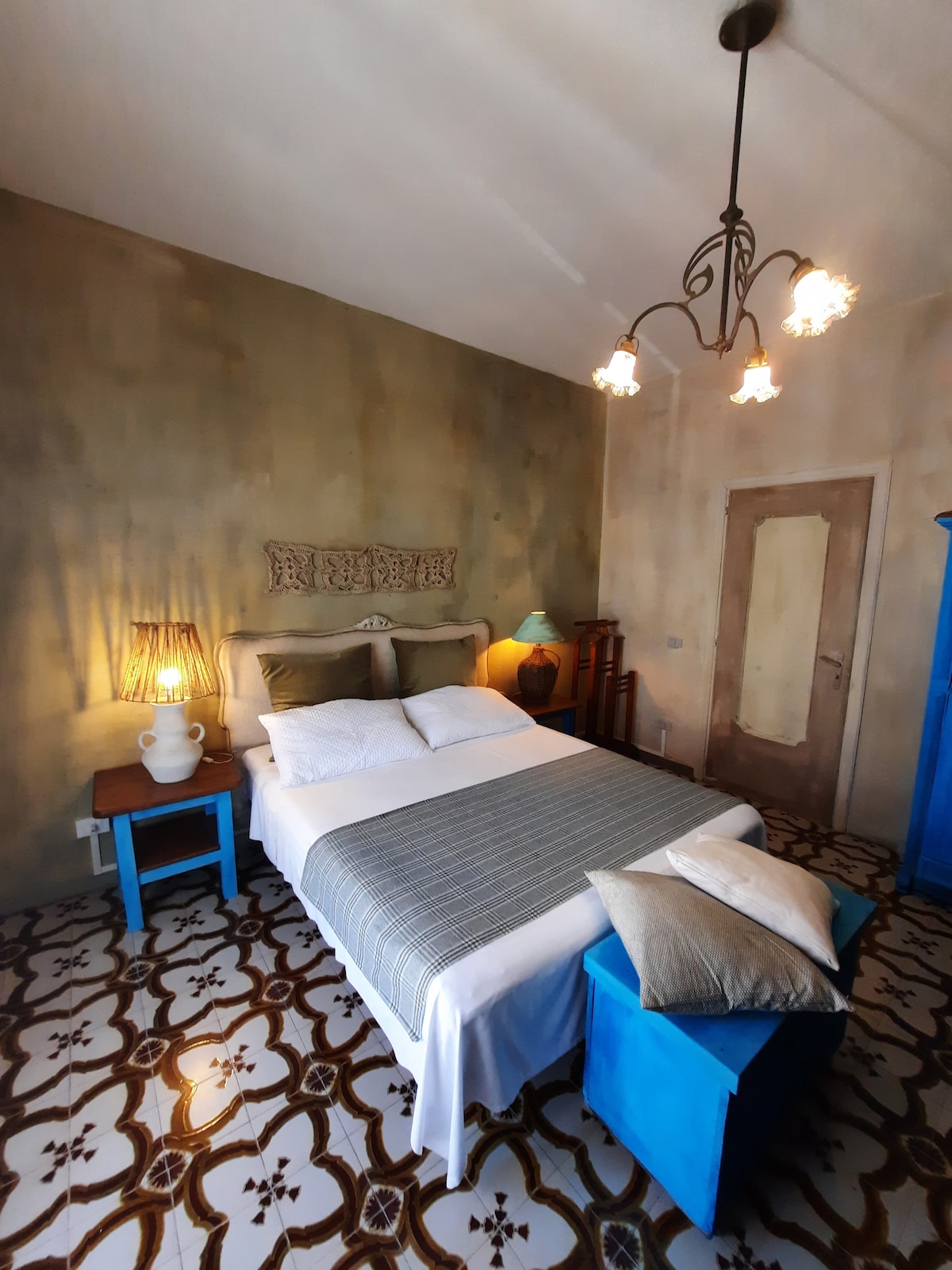 A well-lit bedroom features a spacious bed with crisp white linens and a gray throw. Two bedside tables in contrasting colors flank the bed, each adorned with lamps. Characteristic patterned tiles cover the floor, and unique light fixtures hang from the ceiling.