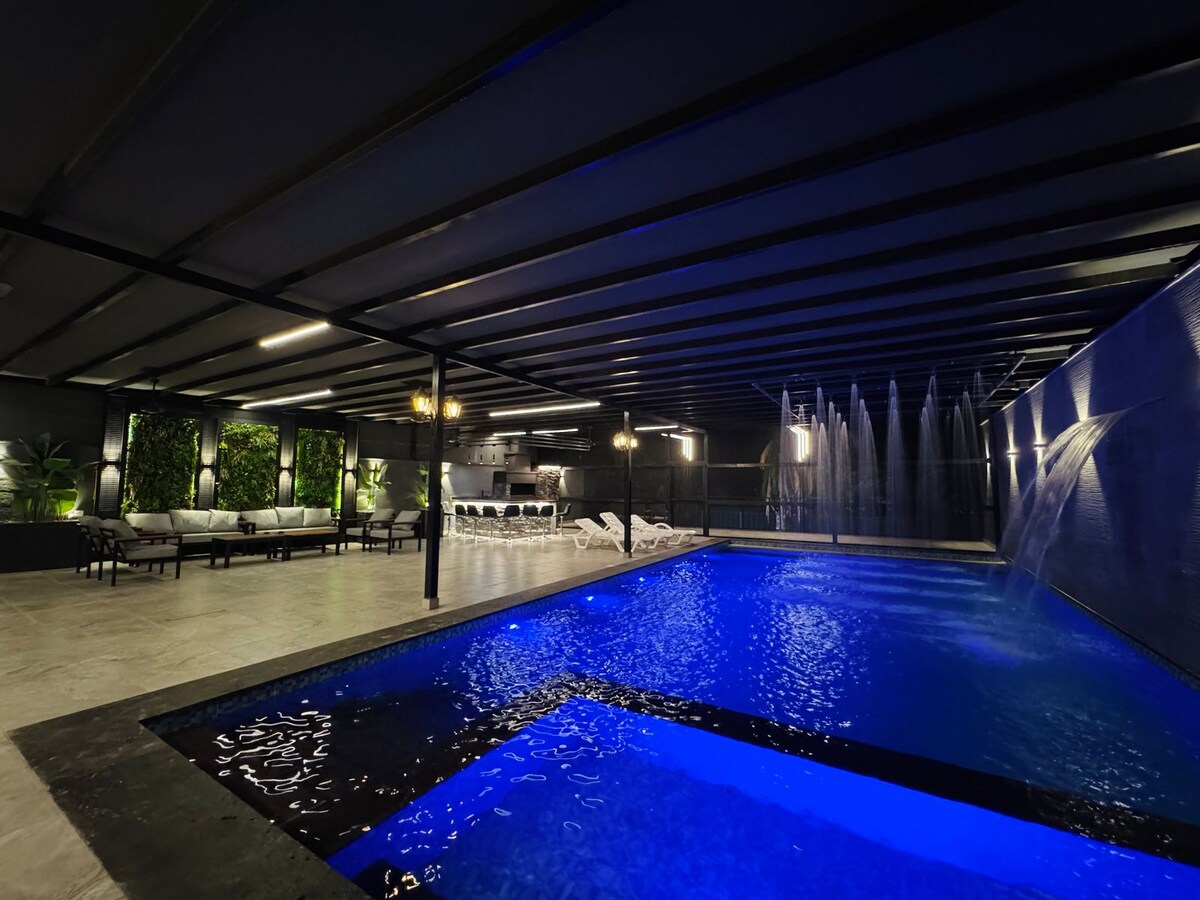 The indoor pool area is illuminated by soft lighting, featuring a vibrant blue swimming pool with a waterfall feature. Comfortable seating is arranged along the sides, surrounded by greenery, creating an inviting atmosphere for relaxation.