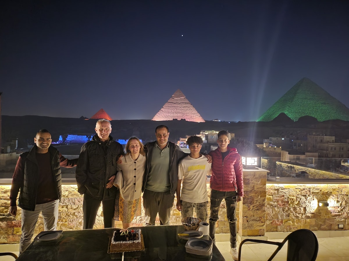 A group of six individuals stands on a balcony, illuminated by soft lighting. The iconic Giza Pyramids are visible in the background, adorned in vibrant colors against the night sky. A table set for dining is positioned in the foreground.