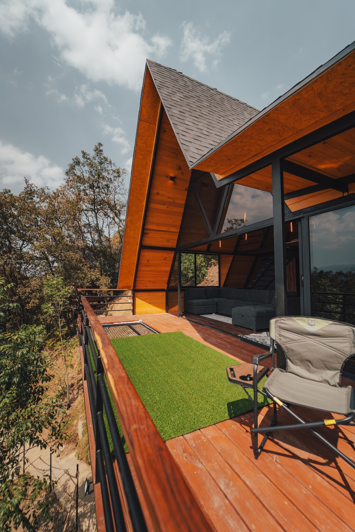 An outdoor deck showcases a blend of wood and glass, featuring a lounge area and a folding chair. The slanted roof adds architectural interest, while greenery frames the view, enhancing the natural surroundings.