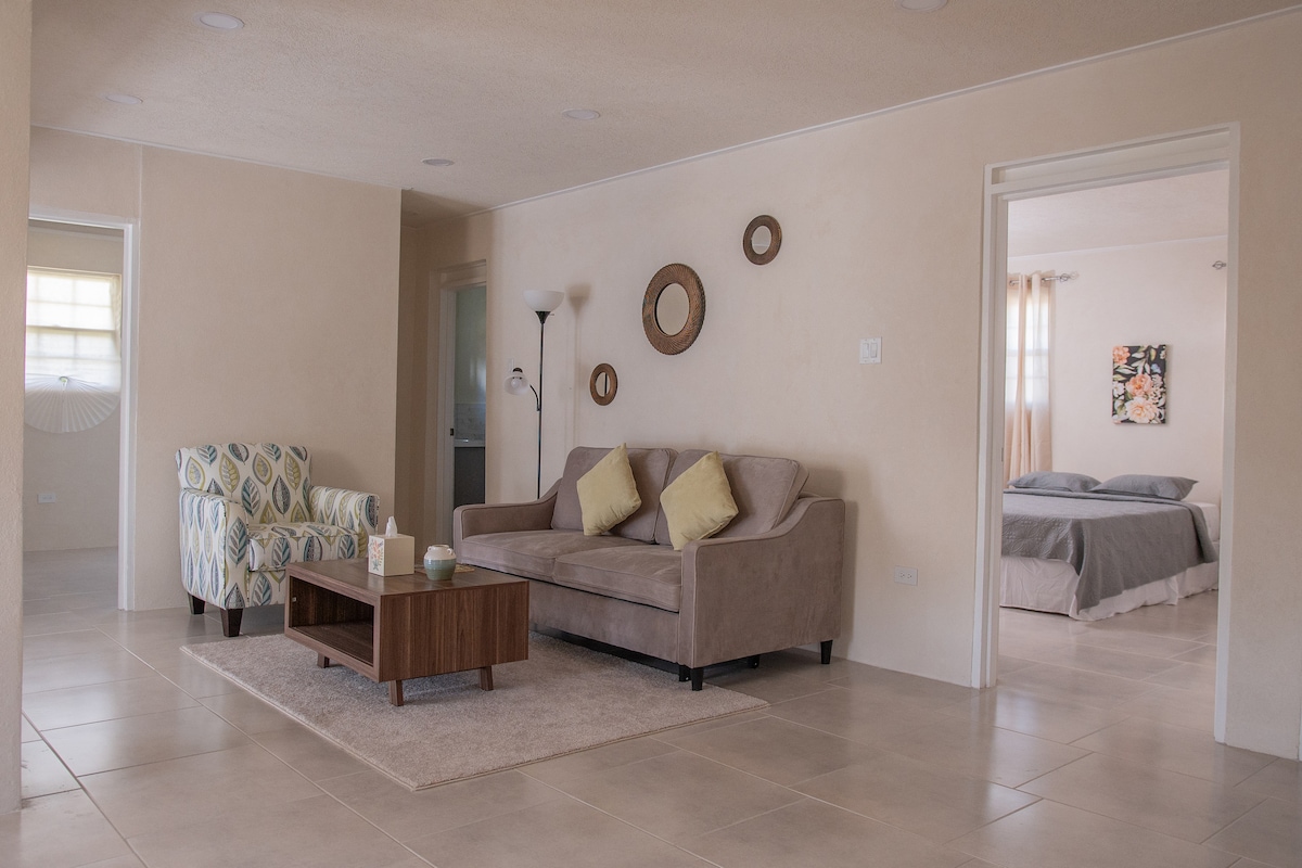 The living area features a comfortable beige sofa paired with two accent pillows, facing a modest coffee table. An accent chair adds a touch of color. Soft lighting reflects off smooth walls, while doorways lead to bedrooms, seamlessly connecting spaces.