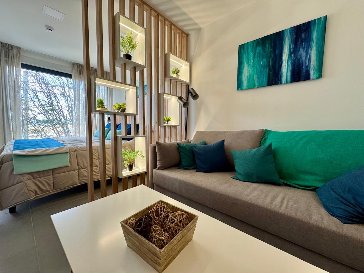 A comfortable living space features a neutral-toned sofa adorned with cushions in teal and dark green. A light wood partition displays potted plants, complementing the artwork above. Natural light filters through a large window, illuminating the area and creating a relaxing ambiance.