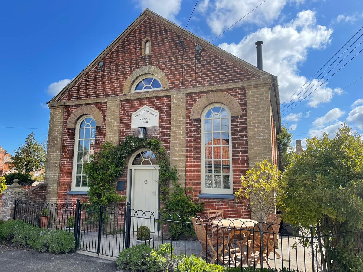 The exterior of a historic chapel is captured, featuring classic brickwork and arched windows. A small outdoor seating area with a table and four chairs is visible, framed by greenery. Clear blue skies with scattered clouds create a bright and inviting atmosphere.