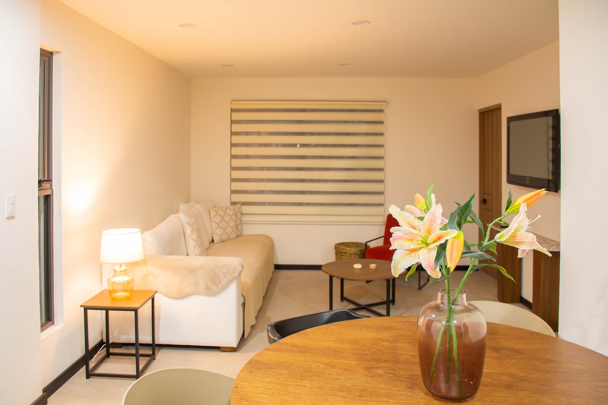 A well-lit living area features a cozy sofa with neutral tones and a stylish coffee table. A round dining table with chairs is visible, accompanied by a glass vase holding fresh flowers. Large windows allow natural light to fill the space.