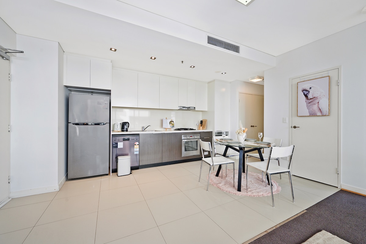 A modern kitchen area is visible, featuring stainless steel appliances, including a refrigerator and oven. A dining table with four chairs is positioned nearby, and a soft rug is placed under the table. Bright lighting enhances the open and airy feel of the space.