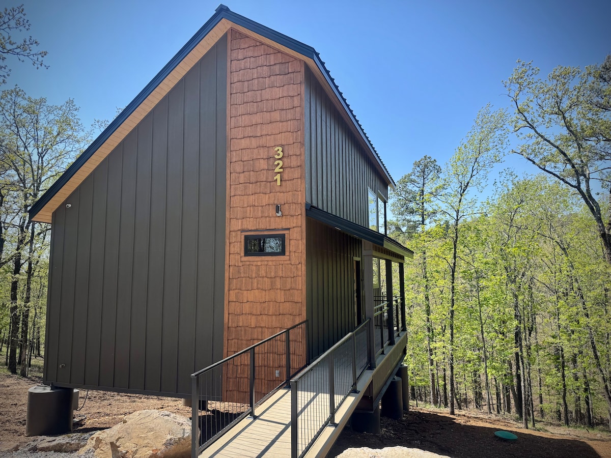The modern cabin features a combination of wood and metal siding, framed by a walkway leading to an entrance. Large windows allow natural light to flood the interior, while surrounding trees create a serene and private atmosphere.