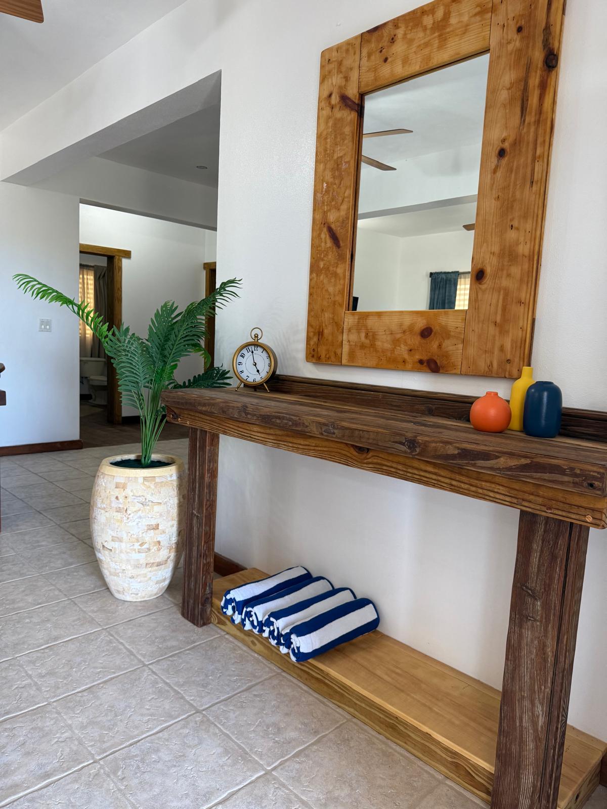 A wooden console table is positioned against a wall, featuring a large mirror above it. A decorative clock and colorful vases are placed on the table. A tall plant is situated in a neutral-colored pot nearby, with neatly rolled towels arranged on the lower shelf.