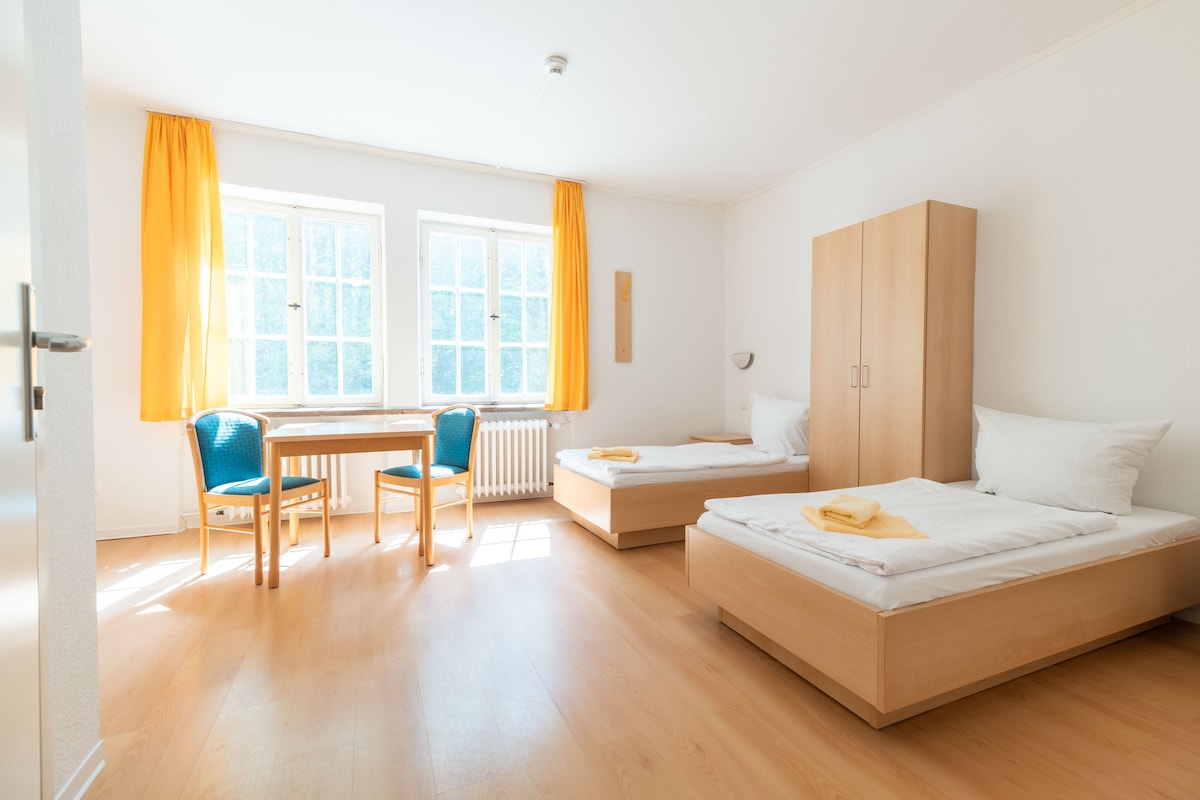 The room features two spacious beds with white linens and pale wood frames. A light wooden table with two blue chairs is positioned near a window with bright yellow curtains, allowing natural light to fill the space. A wardrobe is present along one wall.