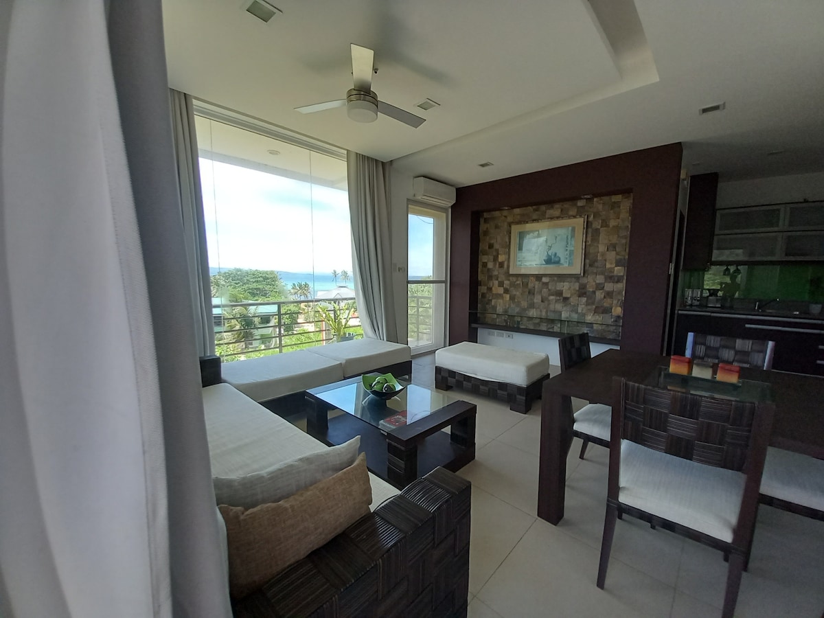 A modern living area features a stylish wooden coffee table centered between two light-colored sofas. Large glass doors frame the ocean view, while soft natural light illuminates the space. A dining table with chairs is visible, creating an inviting atmosphere.