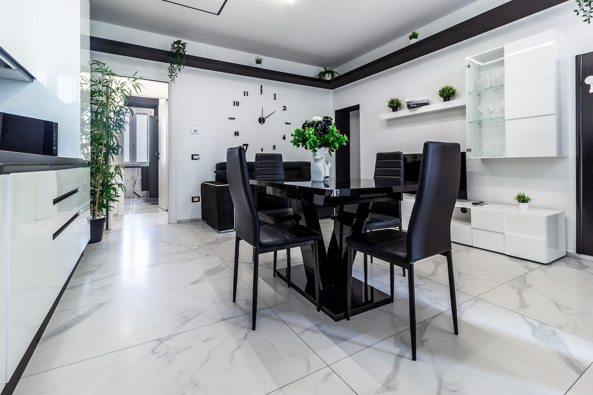 A modern dining area features a black dining table surrounded by sleek black chairs. Light-colored marble flooring reflects natural light, while a wall clock serves as a focal point. A minimalist sideboard and decorative plants add to the contemporary decor.