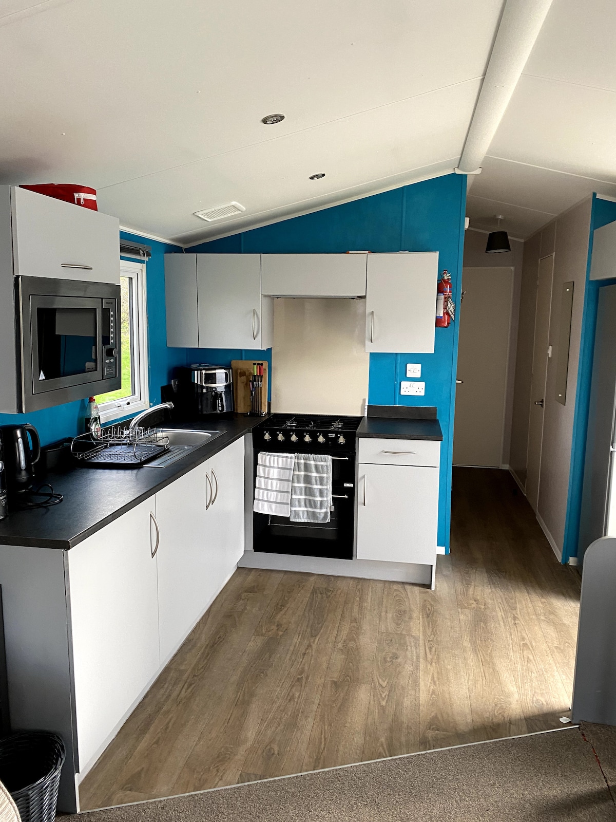 A modern kitchen area features bright blue walls and sleek cabinetry. Essential appliances, including an oven, microwave, and coffee maker, are visible. Counter space is provided for meal preparation, complemented by a window that allows natural light to illuminate the space.