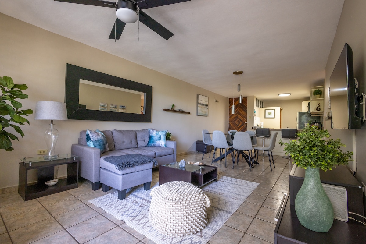 The living area features a comfortable sofa with accent pillows, a stylish coffee table, and a large mirror that enhances the space. A dining table accommodates six guests, while a ceiling fan provides airflow. Natural light enters through the windows, highlighting the neutral décor.
