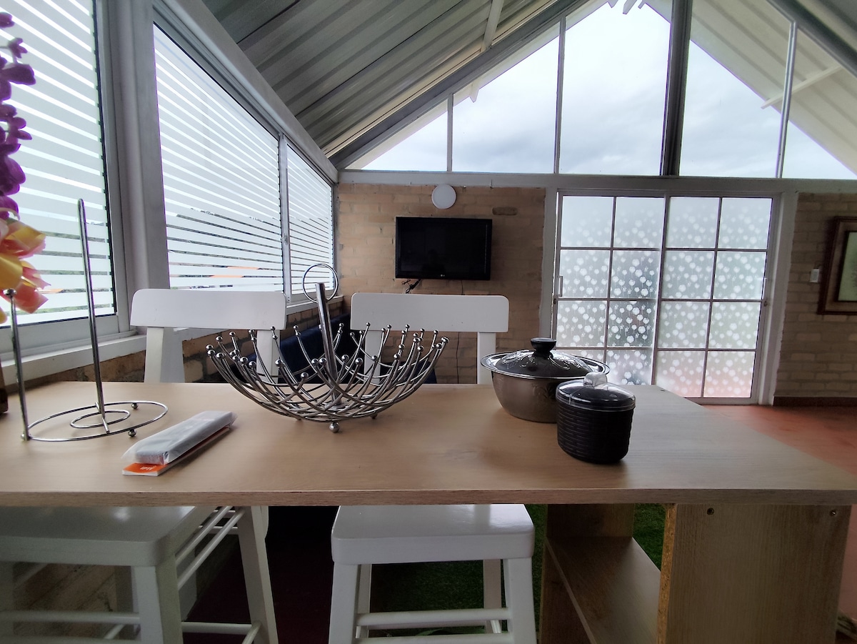 A light-filled dining area showcases a wooden table with sleek white chairs. A decorative bowl is placed on the table, alongside a few cooking vessels. Large windows with modern blinds allow natural light to illuminate the space, creating an airy environment.