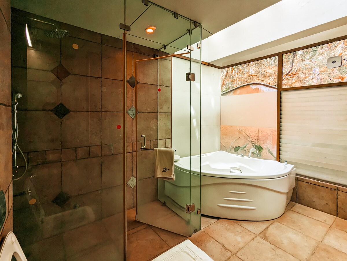 A well-appointed bathroom features a glass-enclosed shower alongside a spacious jacuzzi. Natural light pours in through large windows, creating a bright and airy environment. Neutral-toned tiles extend along the walls and floor, offering a serene setting for relaxation.