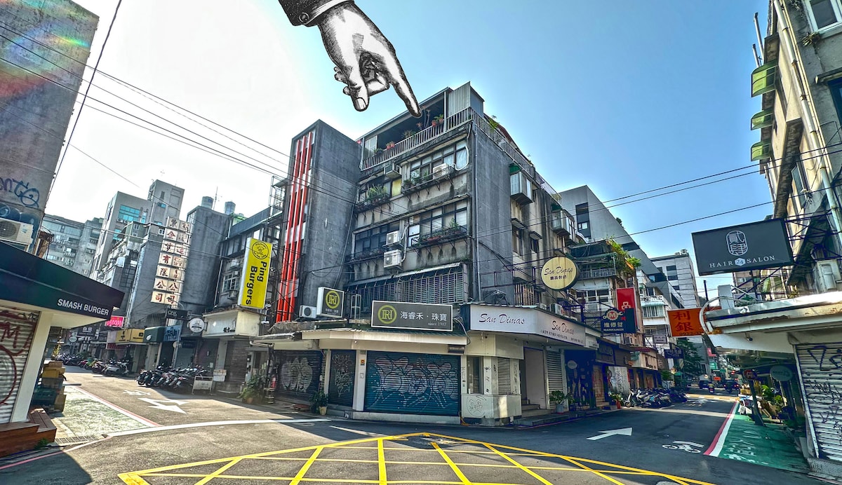A busy urban street scene is captured, showcasing a mix of modern and retro architecture. Colorful signage adorns the building facades, while a clear blue sky is visible above. Streets are lined with shops and restaurants, creating a vibrant atmosphere.