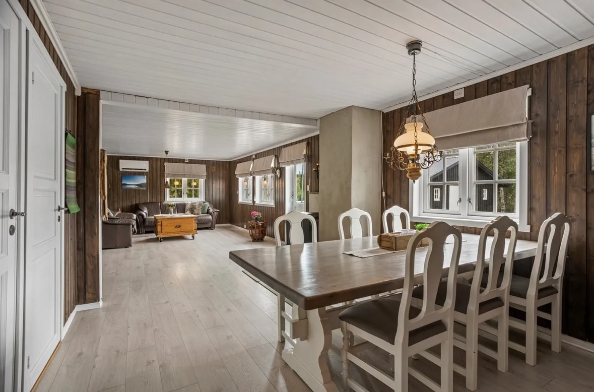 A spacious dining area features a long table surrounded by white chairs, set against a backdrop of wooden walls. Natural light filters through multiple windows, illuminating a connected living space with a cozy seating arrangement in the distance.