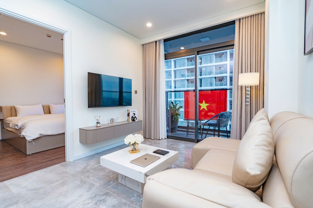 A modern living area features a light-colored sofa facing a flat-screen television mounted on the wall. A coffee table is positioned in front, and large glass doors lead to a balcony displaying a flag. Soft drapes frame the doorway, enhancing natural light.