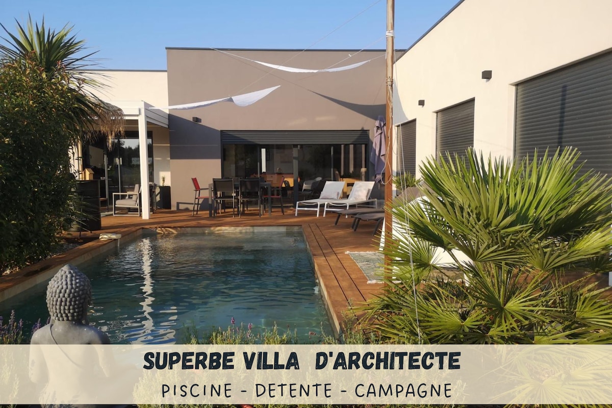 An architect-designed villa is surrounded by lush greenery, featuring a tranquil swimming pool. Decking areas are furnished with loungers and an outdoor dining table, inviting relaxation. A contemporary facade with large windows provides natural lighting and views of the serene outdoor space.