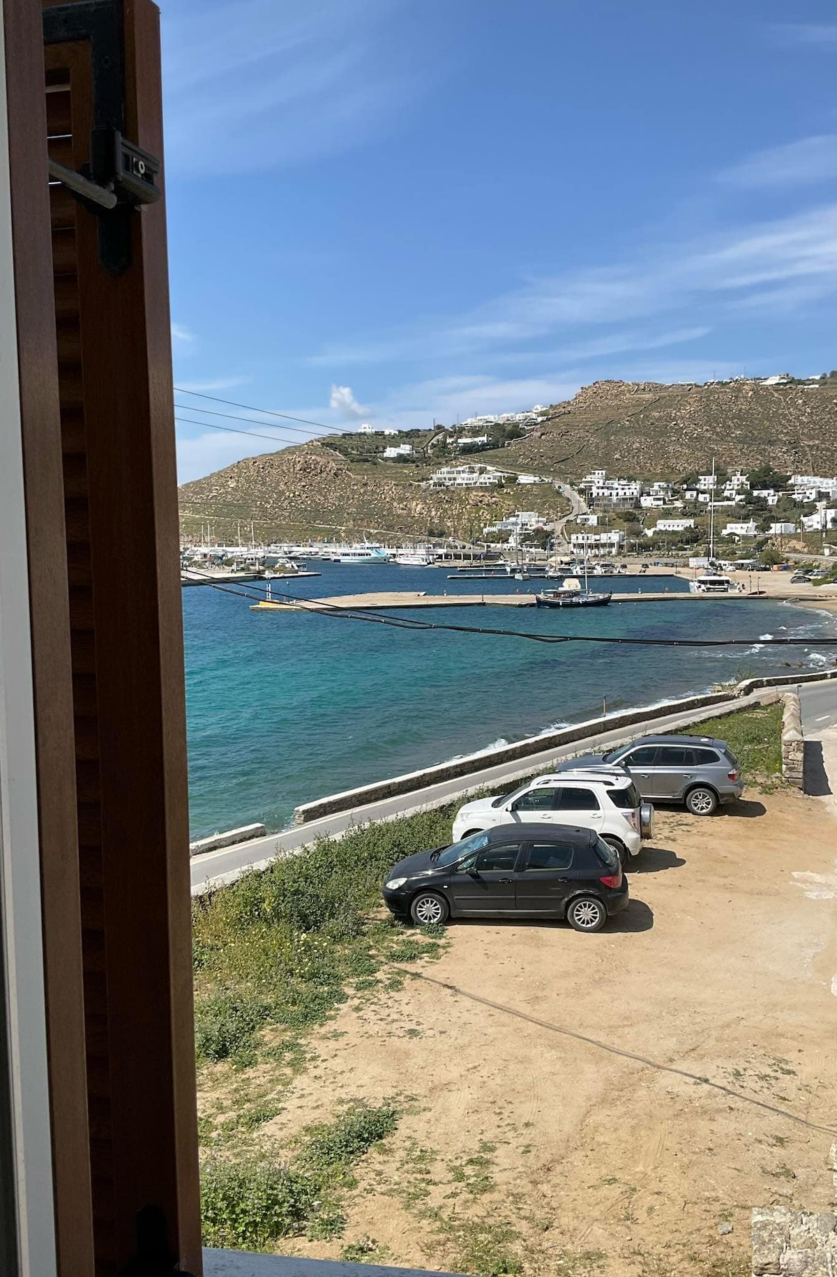 A view of the serene Aegean Sea is captured from an open window. A small harbor is visible, lined with boats and surrounded by gentle hills and white buildings. A parking area with several cars is located in the foreground, leading to a sandy pathway.