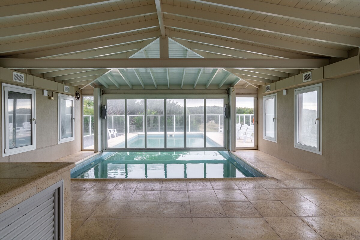 An indoor pool area features large glass windows that provide views of the outdoor landscape. The pool is surrounded by a spacious deck with lounge chairs visible beyond the glass doors, and the ceiling showcases exposed wooden beams.