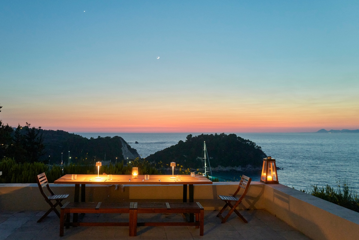 An outdoor terrace is seen at dusk, featuring a long wooden dining table surrounded by five wooden chairs. Soft light from candle lanterns illuminates the table, while a panoramic sea view stretches into the distance, highlighting the transition of colors in the sky.