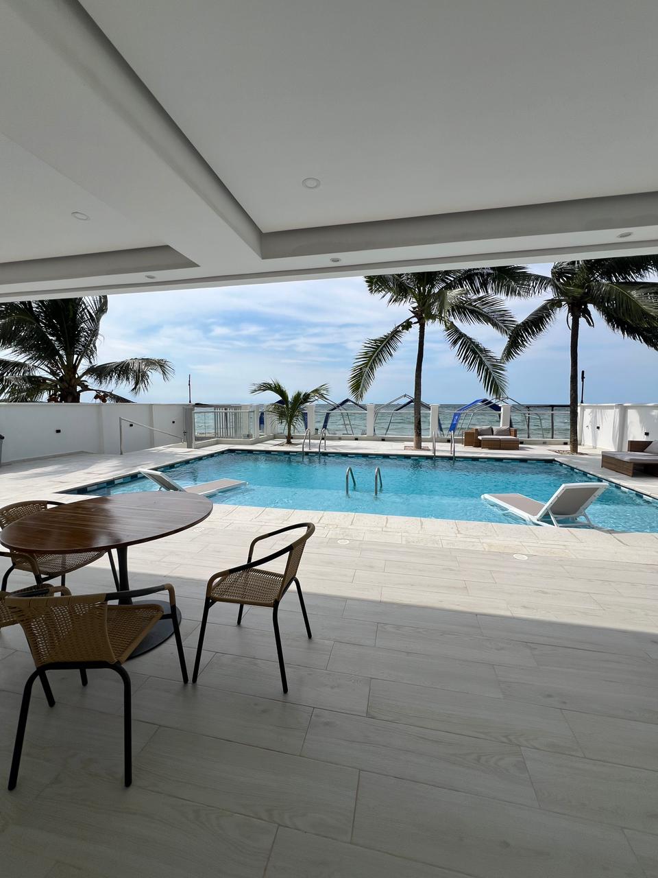 An outdoor area features a swimming pool surrounded by lounge chairs. A round dining table with four chairs sits under a covered terrace, while palm trees gently sway nearby. The ocean is visible in the background, enhancing the outdoor space's inviting ambiance.