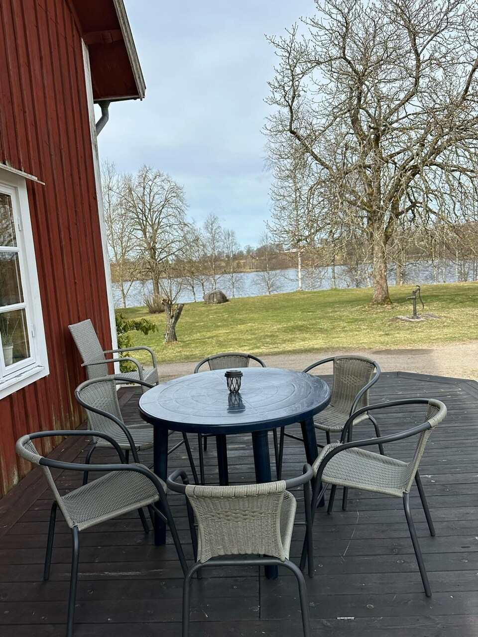 A round table with six chairs is positioned on a wooden deck. In the background, a serene lake and trees can be seen, creating a peaceful outdoor setting. The scene captures the essence of relaxation and gathering by nature.