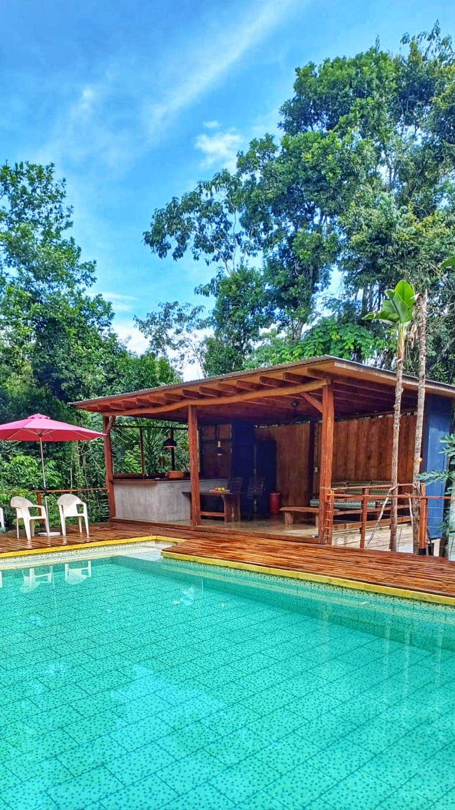 A wooden deck surrounds the tropical pool, offering a space for relaxation. A shaded seating area is visible under a roof, furnished with a table and benches. Nearby, a red umbrella provides additional shade, while lush greenery creates a serene atmosphere.