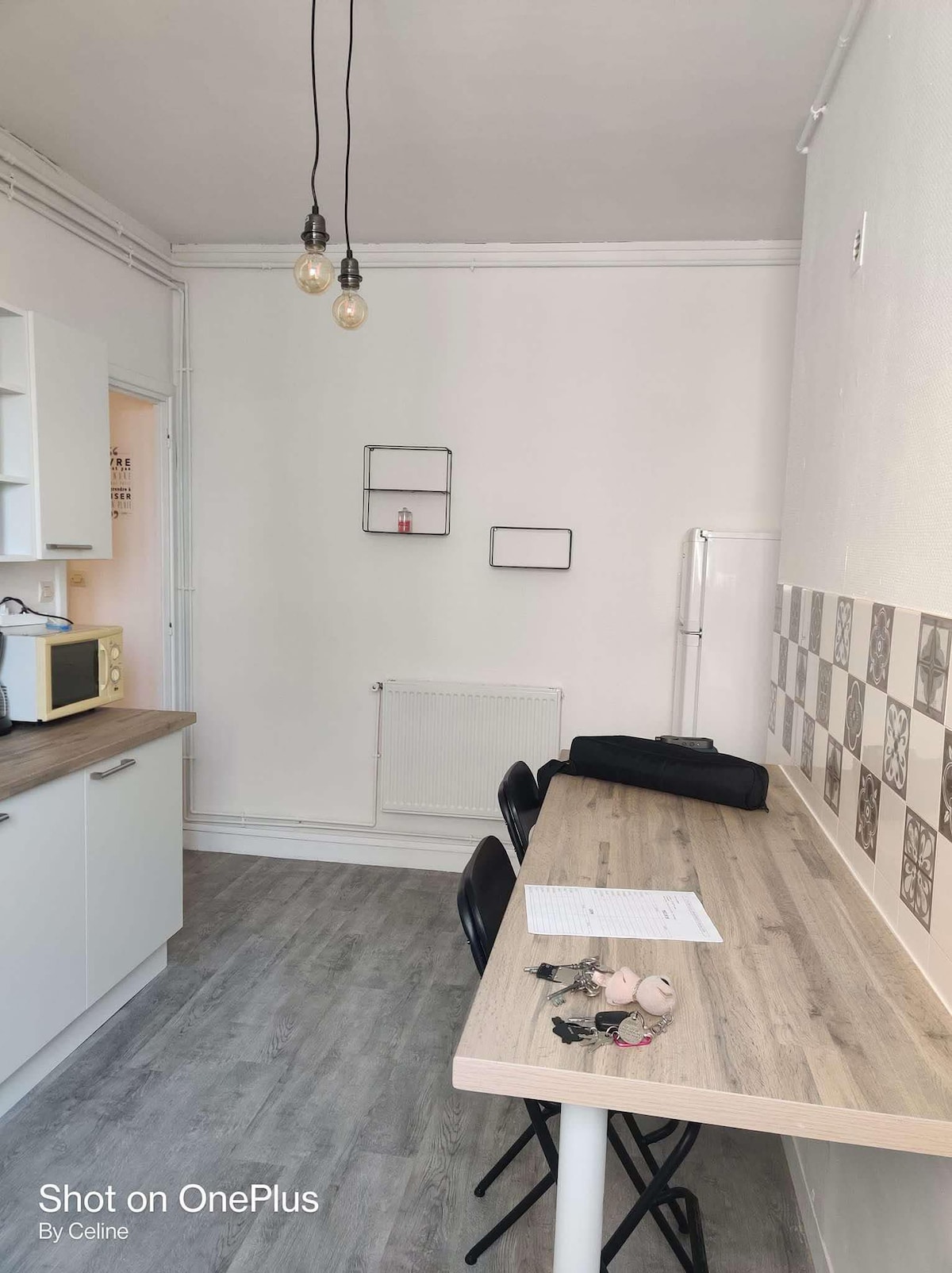 A bright kitchen area features a light wooden table with seating for four. A wall-mounted radiator is positioned beside the table, and a microwave is located on the countertop between cabinets. Two pendant lights hang from the ceiling, illuminating the space.